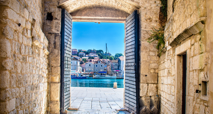Doorkijkje door een stenen stadspoort in Trogir naar de gezellige riva en haven
